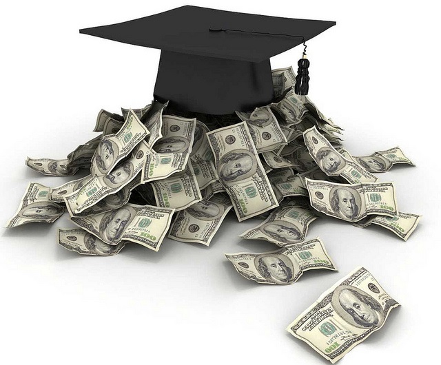 A black graduation cap sits on a pile of 100 dollar bills.
