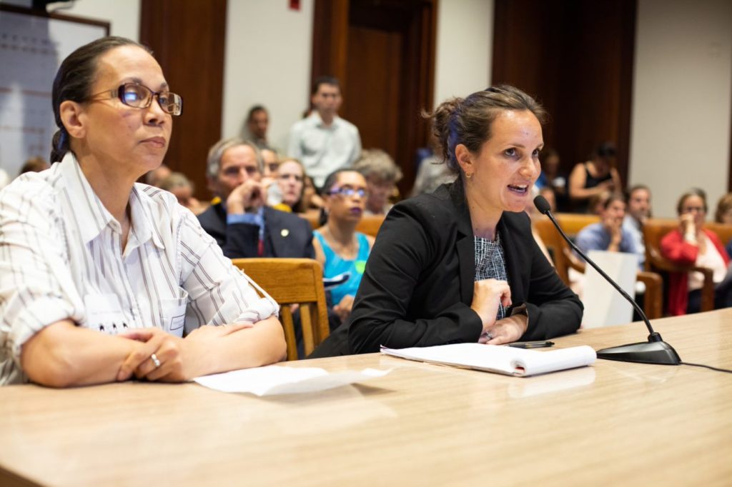 Nicole Summers (right) sits at a table in court, speaking into a microphone to testify in front of the Joint Committee of the Judiciary. Andrea Nickerson (left) is seated next to her.