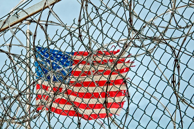 A tattered American flag is seen through prison gates covered in barbed wire