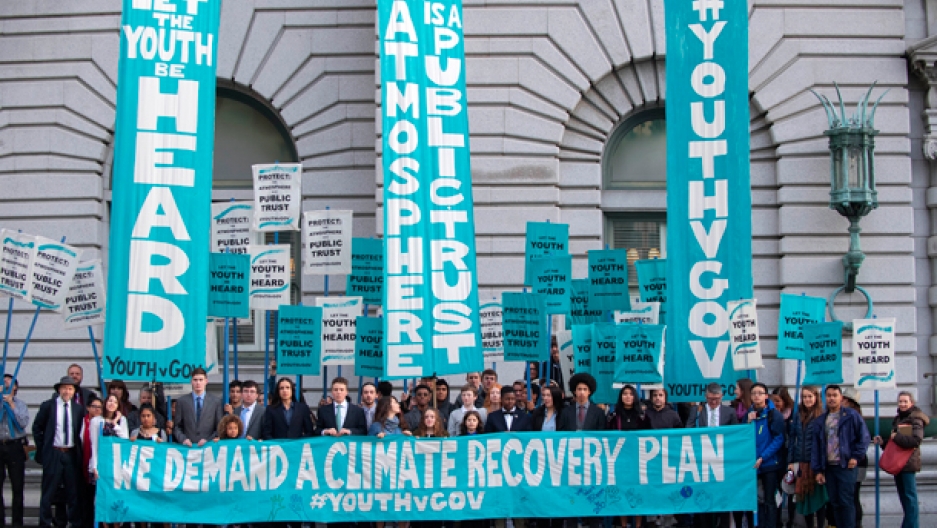 Youth stand in front of Court of Appeals holding a teal banner reading 'We demand a climate recovery plan #YouthvGov' in white text. Other vertical teal colored banners read: 'Let the youth be heard', 'Atmosphere is a public trust', and '#YouthvGov'. Youths in the background hold a variety of white and teal pickets,