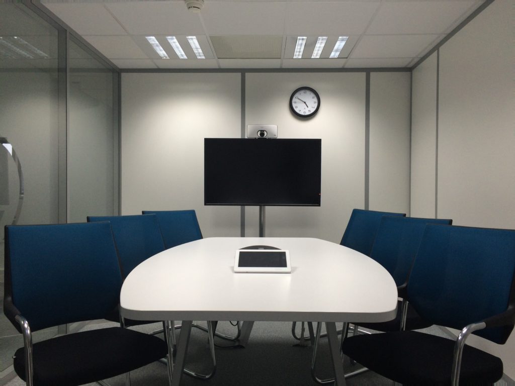 A white conference room with one glass wall to the right and a white table in the middle. The table is surrounded by six blue chairs, three on each side, facing the front of the room. A large black TV is mounted on the back wall.