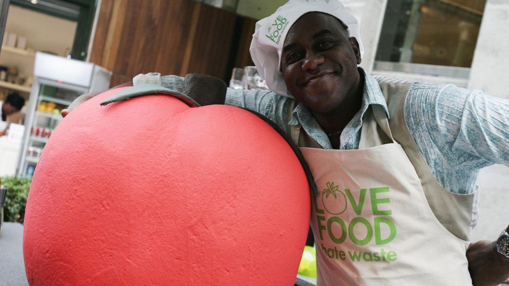 Ainsley Harriot smiles at the camera during the "Love Food, Hate Waste" event, with one arm around a large styrofoam red apple