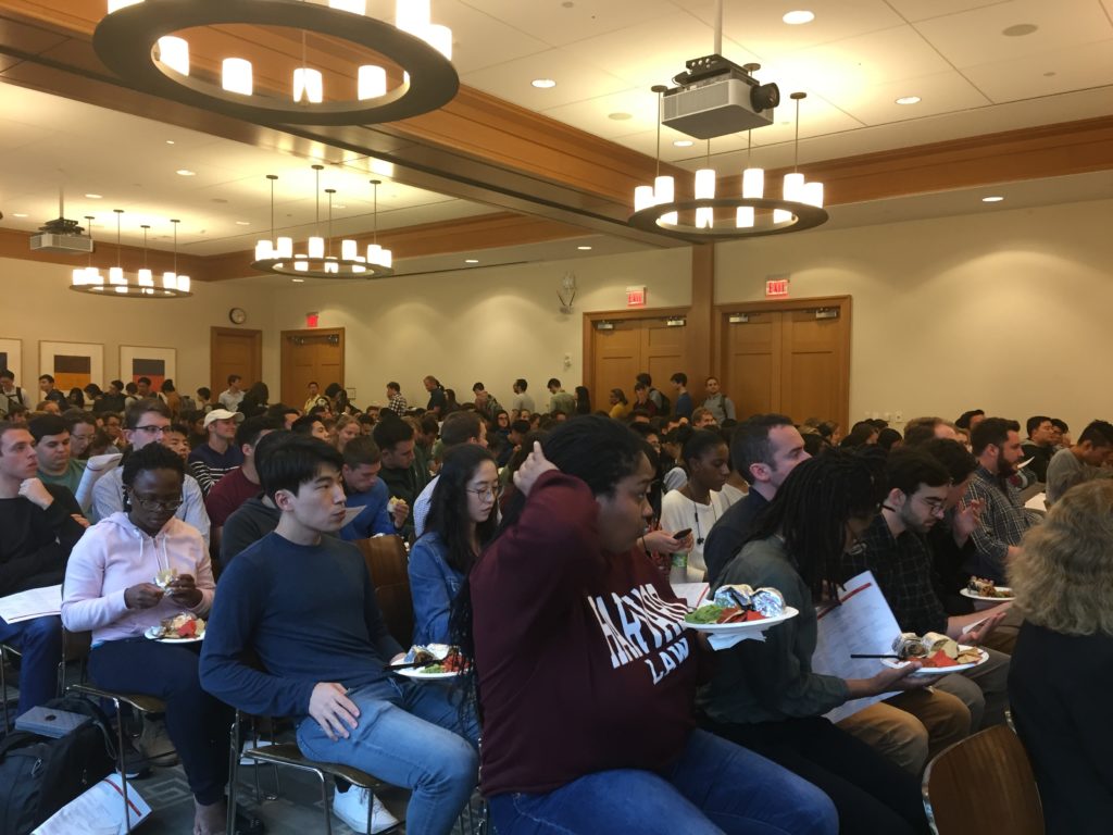 Group of students sit in large panel room with plates of food.