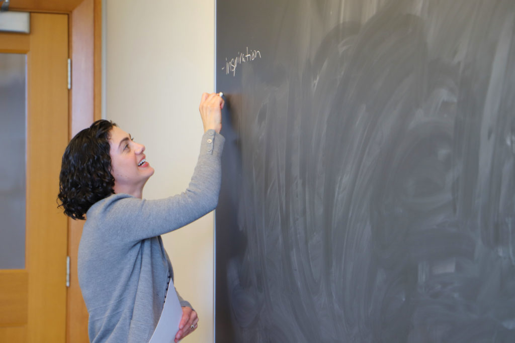 Susan Farbstein stands in front of a black board, writing the word 'inspiration' in white chalk