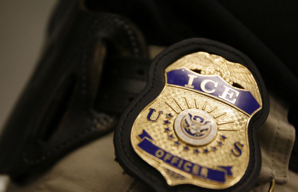 An ICE officer badge in gold and blue is shown clippped to a belt of an officer